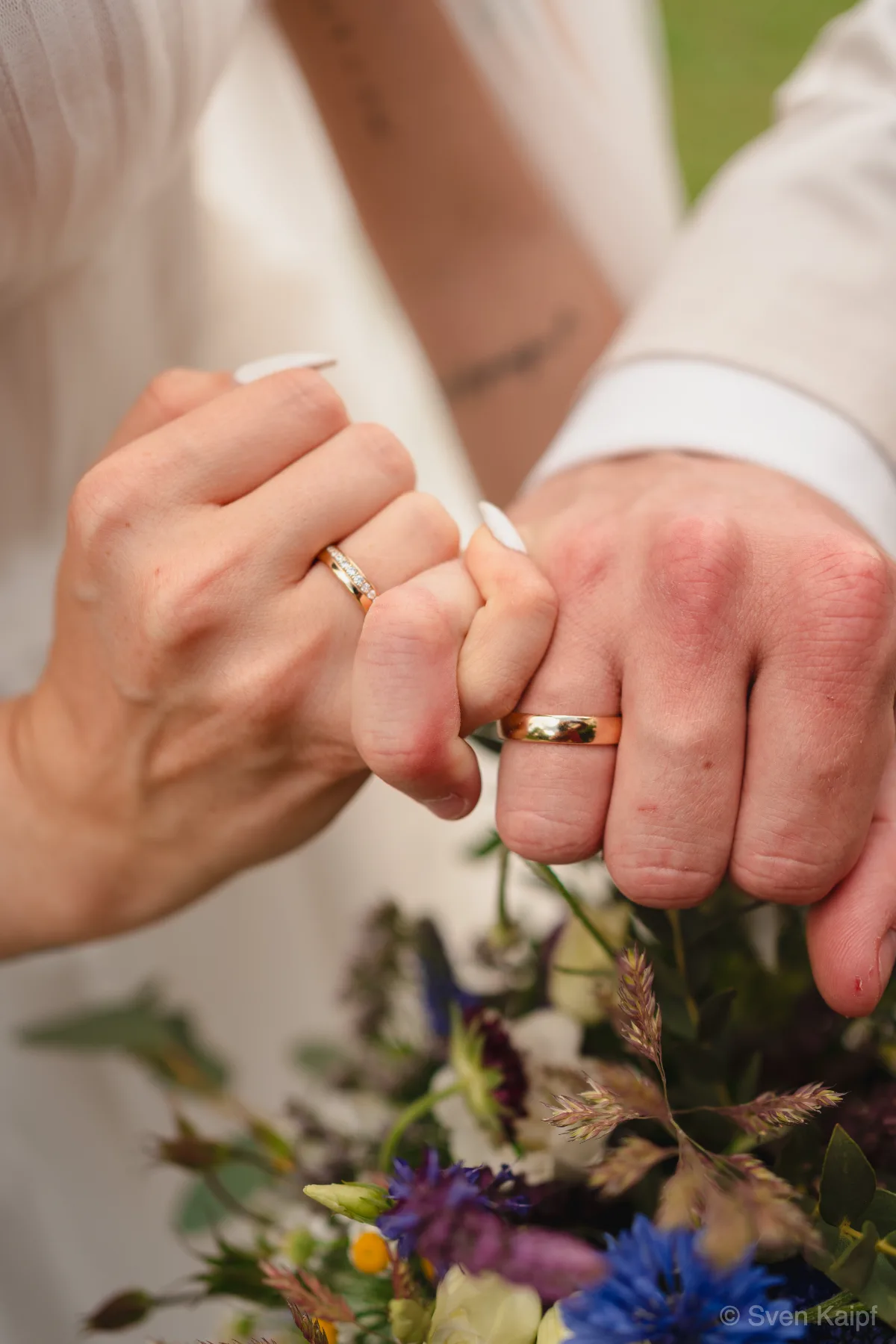 Hochzeit Detailaufnahme: Eheringe an den Händen des Hochzeitspaares, kleine Finger ineinander gehakt, davor ein Blumenstrauß
