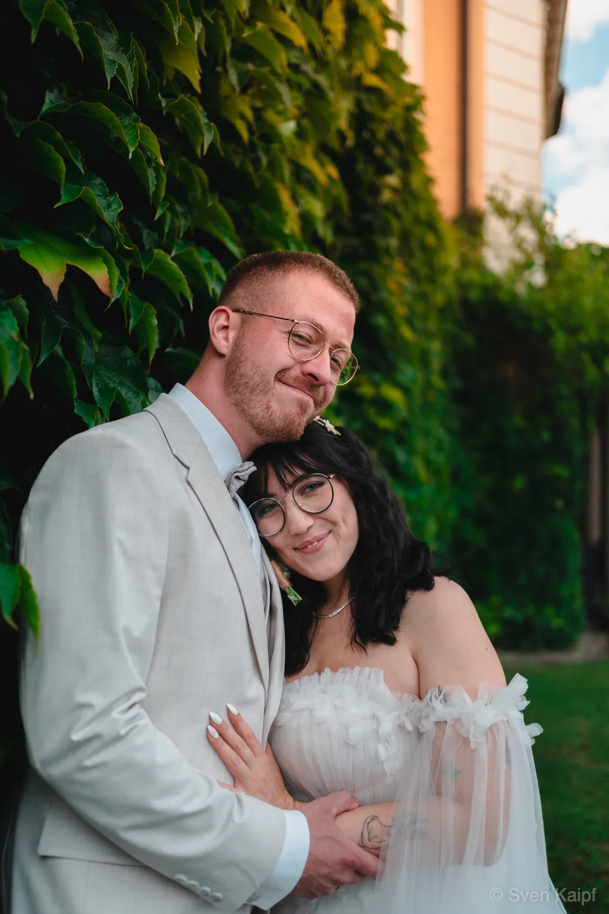 Hochzeitsportrait vor einer Efeuwand: Hochzeitspaar lehnt die Köpfe aneinander und lächelt in die Kamera