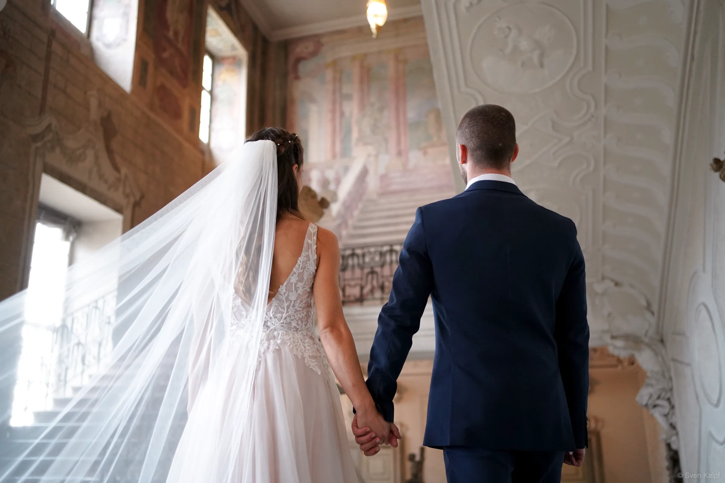 Hochzeitsfoto im Treppenhaus: Hochzeitspaar geht Hand in Hand die Treppe hinauf, von hinten fotografiert, Schleier weht