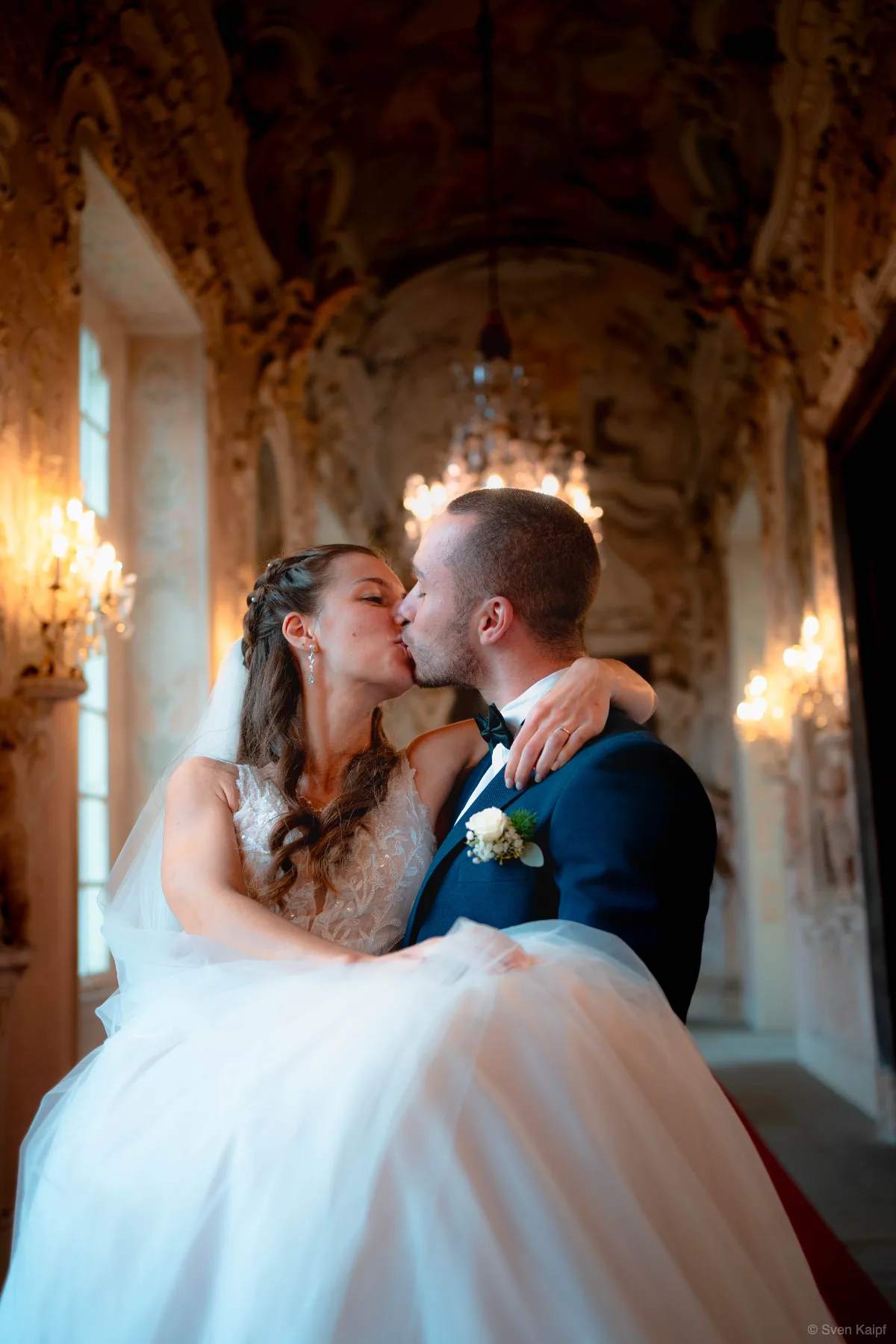 Hochzeitspaar küsst sich in einem historischen Gang mit Kronleuchtern, Bräutigam trägt die Braut auf den Armen