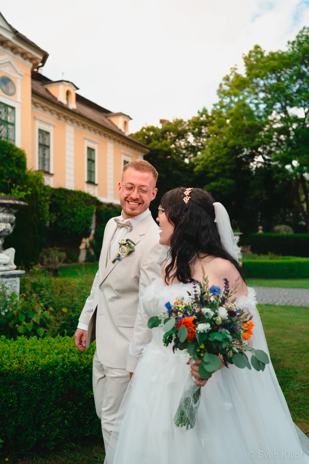 Hochzeitspaar geht lachend durch einen Garten vor einem gelben Barockschloss, Braut mit Blumenstrauß – Hochzeitsbild im Schlossgarten